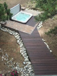 A wooden walkway leading to pavers, and wood deck, and hot tub